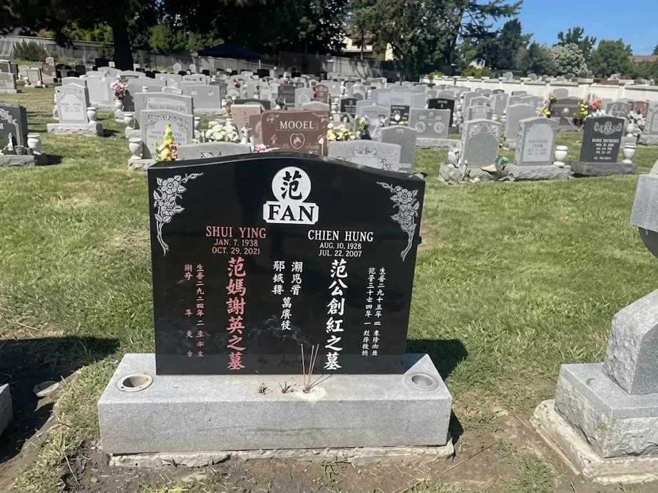 Black granite upright headstone with English and Chinese lettering and carved floral corners in a cemetery