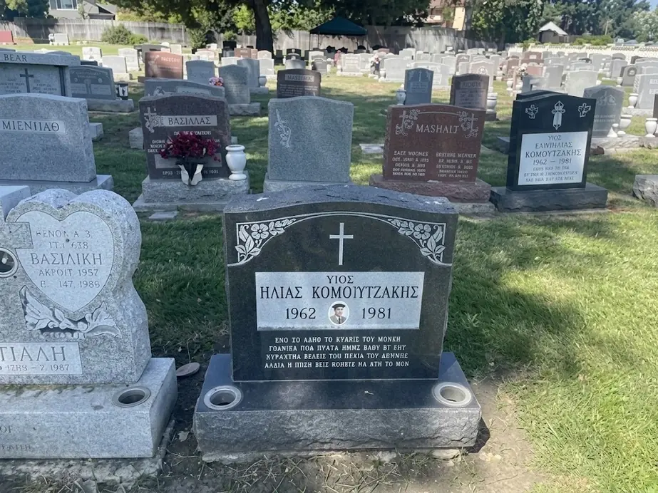 Black granite upright with a cross, carved floral border and Greek lettering in a cemetery